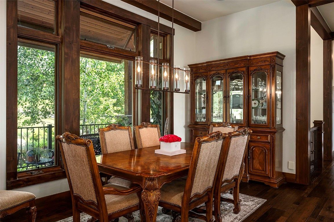 This is an interior shot of a formal dining room featuring a large wooden table with upholstered chairs. A modern chandelier hangs above the table, and a wooden china cabinet stands against the wall. Large windows offer a view of the outdoors, and the room is decorated in a traditional style with dark wood accents.