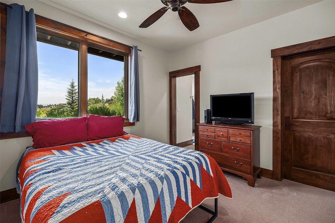 This is a cozy bedroom featuring a bed with a red, white, and blue quilt, red pillows, and a window with blue curtains. A wooden dresser with a television on top sits against the wall, and a wooden door is visible to the right. The room has a warm and inviting feel, with natural light streaming in from the window.