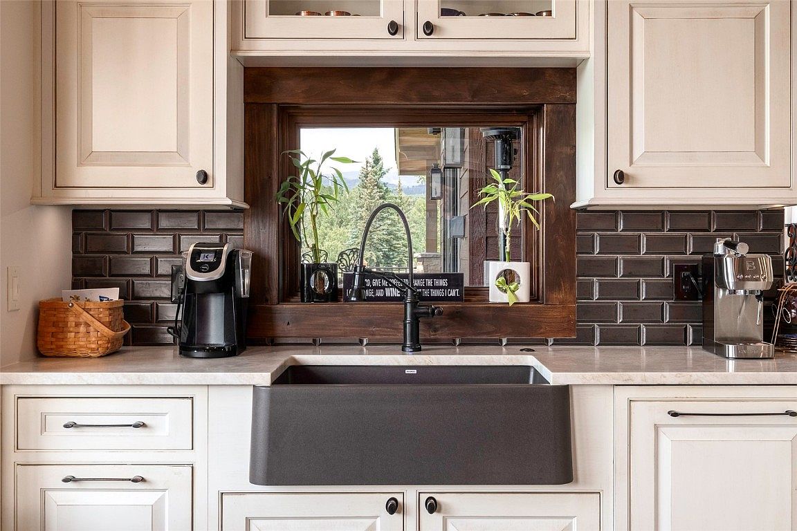This image showcases a well-appointed kitchen area, focusing on the sink and surrounding countertop space. The kitchen features white cabinetry with dark hardware, a dark farmhouse-style sink, and a dark faucet. A window above the sink provides natural light and a view of the outdoors, while decorative plants and a sign add a touch of personality.