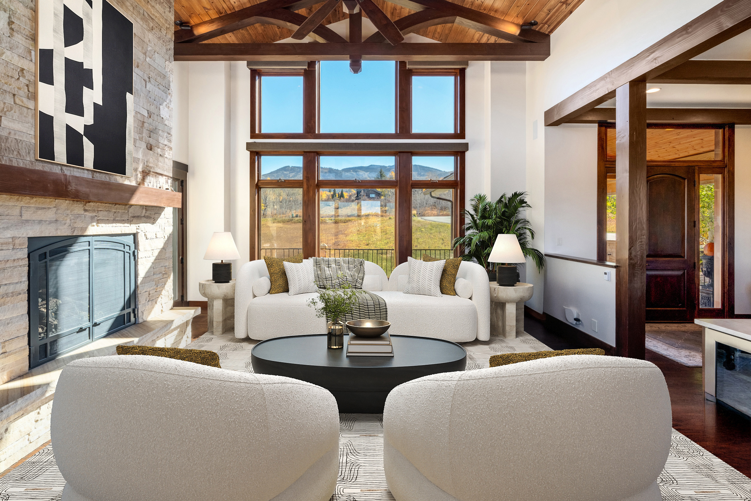 The living room features a modern yet rustic design, with a stone fireplace, wooden ceiling beams, and large windows showcasing a mountain view. The neutral color palette and plush furnishings create a cozy and inviting atmosphere. A black coffee table is centered between the seating areas adds a touch of sophistication.