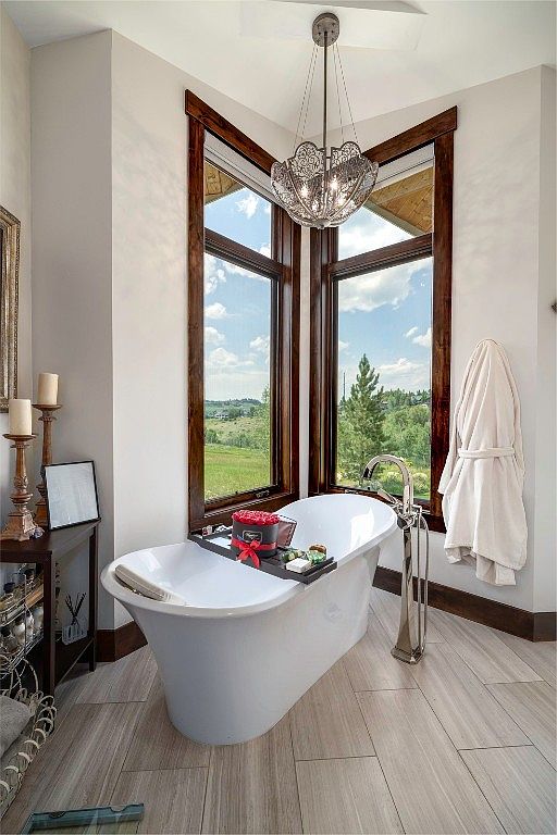 This is a luxurious primary bathroom featuring a freestanding white bathtub positioned beneath a corner window with a scenic view of rolling hills. The room is illuminated by an ornate chandelier, and a plush white robe hangs on the wall. The flooring consists of light-colored wood-look tiles, and the overall aesthetic is elegant and serene.