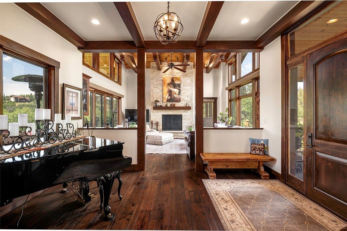 This is an interior shot of a luxurious living room featuring dark hardwood floors, exposed wooden beams, and a stone fireplace. A grand piano sits to the left, while a cozy seating area is visible through the opening, creating an inviting and elegant atmosphere. The room is well-lit with natural light and recessed lighting, enhancing the warm and sophisticated ambiance.
