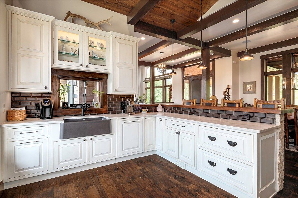 This is a well-lit kitchen featuring white cabinetry, dark brown subway tile backsplash, and a dark farmhouse sink. The kitchen has a rustic-modern style with wooden beams on the ceiling and dark wood flooring. The countertops are light-colored, and the hardware is dark, creating a nice contrast.
