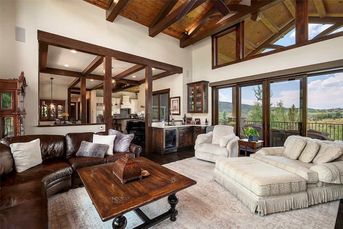 This is an interior shot of a luxurious living room featuring a brown leather sectional sofa, a light-colored chaise lounge, and a rustic wooden coffee table. The room boasts high ceilings with exposed wooden beams and large windows offering a view of the outdoors. A partial view of the kitchen is visible through a wooden frame, adding depth to the space.