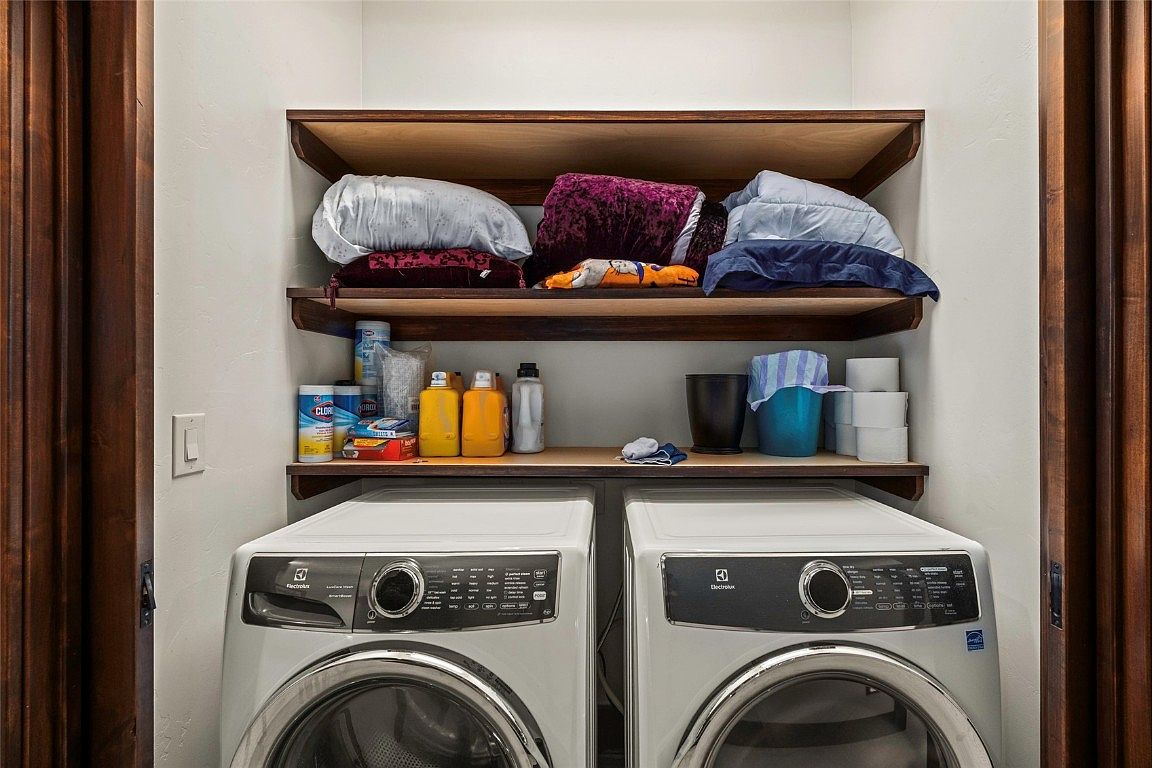 This is an interior shot of a laundry room featuring two front-loading Electrolux washing machines. Above the machines are two wooden shelves holding laundry supplies, including detergent, bleach, and folded items. The room is flanked by dark wood door frames, creating a clean and organized space.