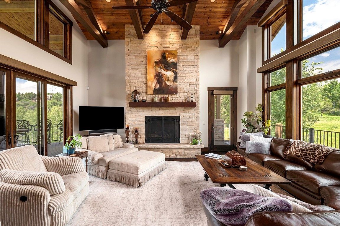 This is an interior shot of a luxurious living room featuring a stone fireplace as a focal point, complemented by exposed wooden beams on the ceiling. The room is furnished with comfortable seating, including a plush armchair and a leather sofa, arranged around a wooden coffee table. Large windows provide ample natural light and views of the outdoor landscape, enhancing the room's inviting and upscale ambiance.