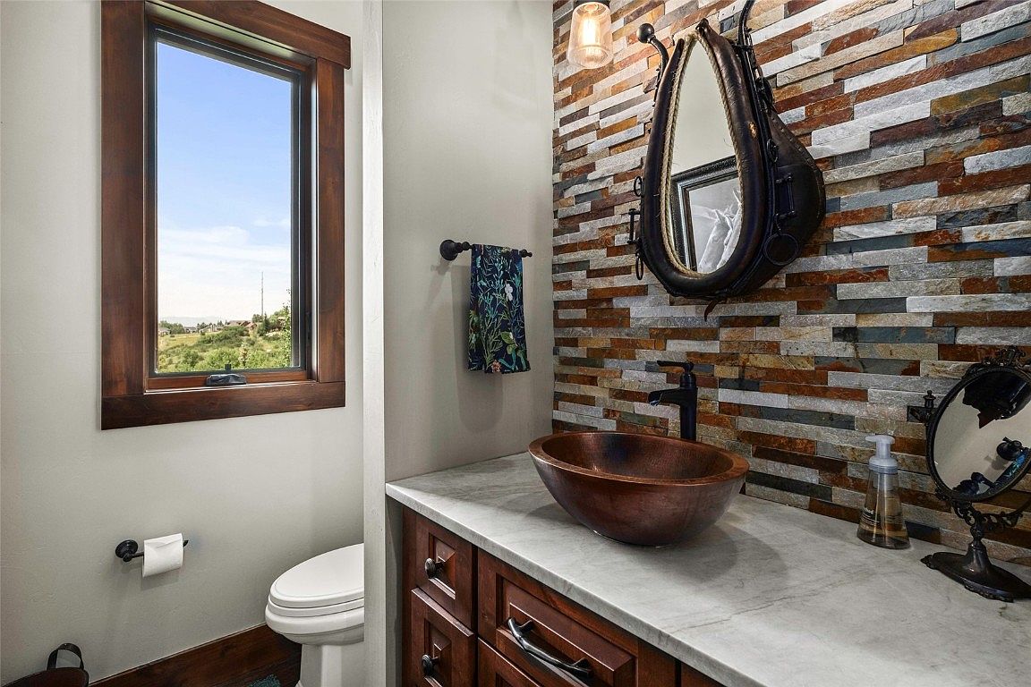 This is a well-appointed guest bathroom featuring a unique copper vessel sink and a decorative stone accent wall. The vanity is made of dark wood with a light countertop, and an interesting mirror adds character. A window provides natural light, and the overall design is stylish and inviting.