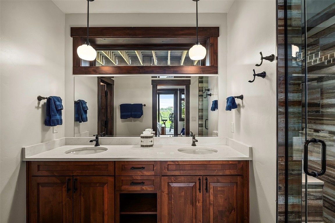 This is a primary bathroom featuring a double vanity with a white countertop and dark wood cabinetry. Above the vanity is a large mirror with a dark wood frame, and pendant lights hang on either side. A glass-enclosed shower is visible to the right, showcasing wood-look tile, creating a spa-like atmosphere.