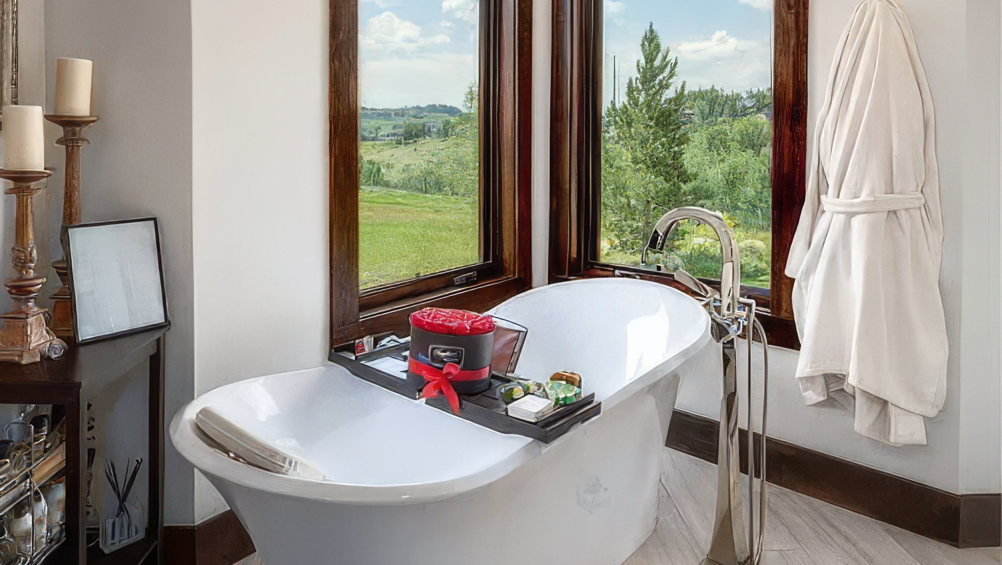 This is a luxurious primary bathroom featuring a standalone soaking tub next to a large window with a view of the outdoors. The room is well-lit and styled with curated bath accessories, creating a spa-like atmosphere. The color palette is neutral and calming, enhancing the sense of relaxation and refinement perfect for a high-end real estate listing.