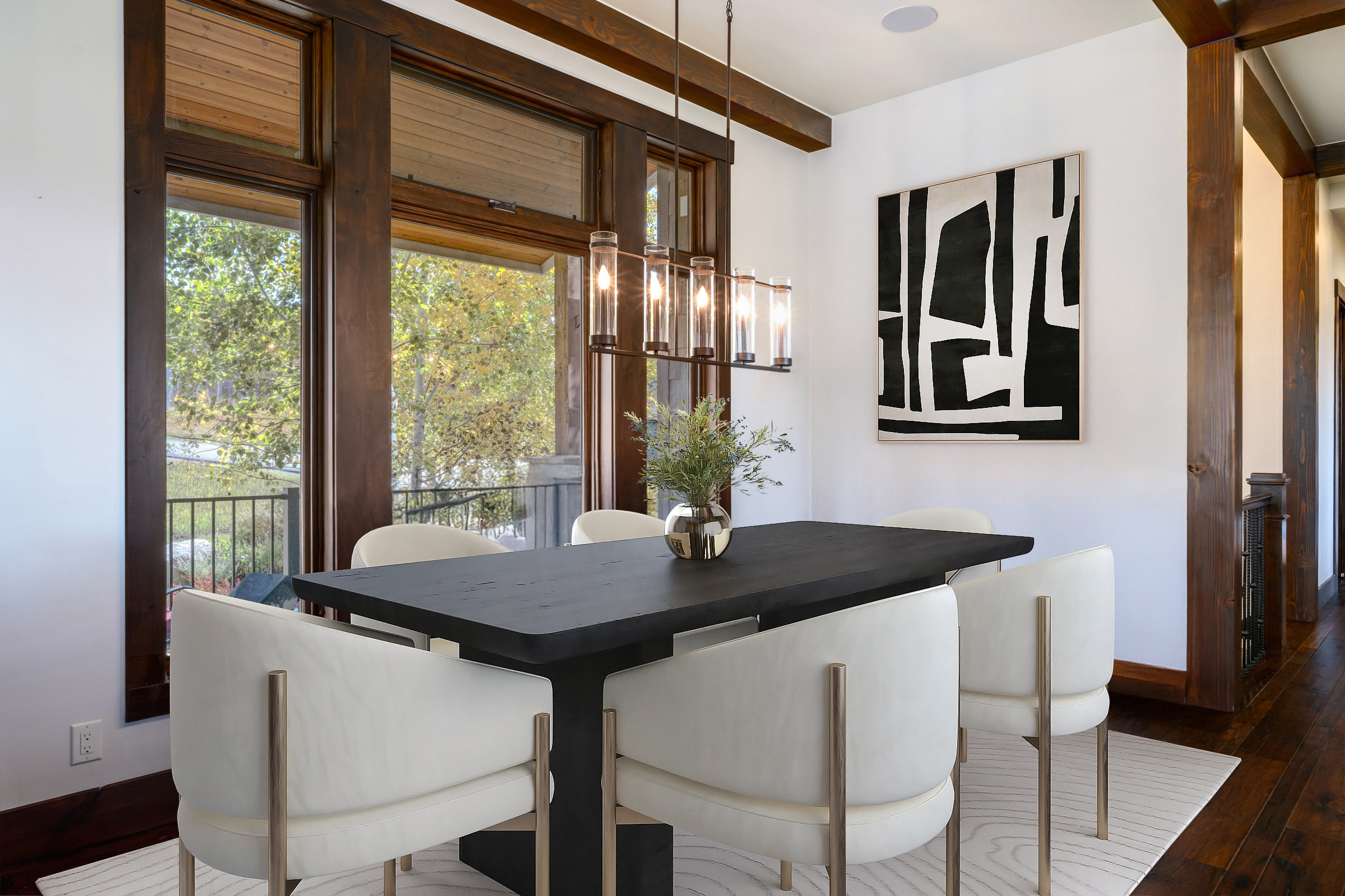This interior shot showcases an elegant dining room featuring a dark wood table surrounded by stylish white chairs with gold accents. A modern chandelier hangs above the table, and an abstract painting adorns the wall. The room boasts a sophisticated atmosphere, enhanced by the natural light streaming through large windows that overlook an outdoor view, making it a perfect space for entertaining.