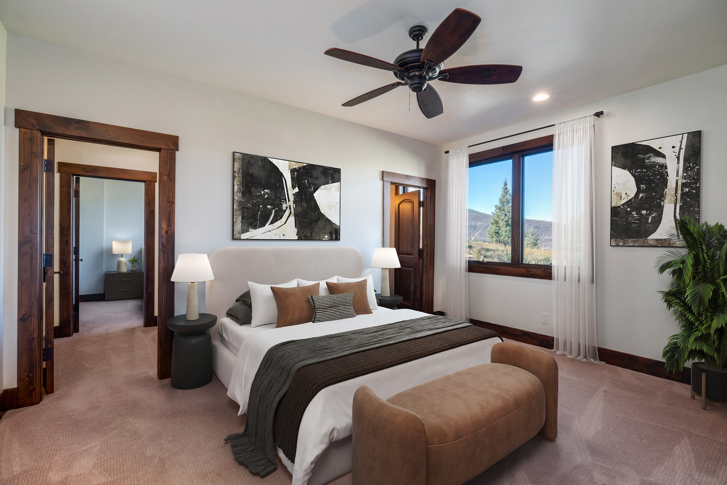 This is a well-lit primary bedroom featuring a king-size bed with neutral-toned linens and pillows, flanked by bedside tables with lamps. The room has a ceiling fan, artwork, and a view of the outdoors through a window. The design presents a comfortable and stylish space for relaxation.
