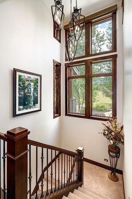 This interior shot showcases a well-lit hallway and staircase. The staircase features dark wood railings and balusters, leading upwards on carpeted steps. A framed picture hangs on the wall, and natural light streams in through large windows, complemented by decorative pendant lights, creating a warm and inviting atmosphere.