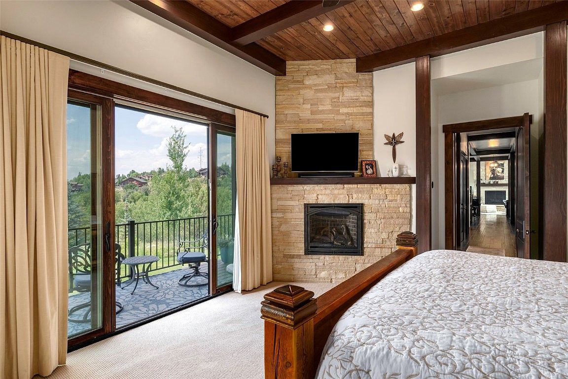This is a primary bedroom featuring a stone fireplace with a mounted television above it. The room has a balcony with outdoor seating, accessible through sliding glass doors. The ceiling is wood paneled with exposed beams, and the room is furnished with a large bed.
