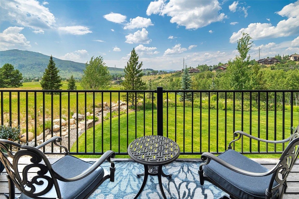 This image showcases a charming patio or deck area, furnished with two ornate chairs and a small, patterned table. The space overlooks a lush green lawn and a scenic landscape with trees and a distant mountain range, all framed by a black metal railing. The overall impression is one of tranquility and outdoor living.