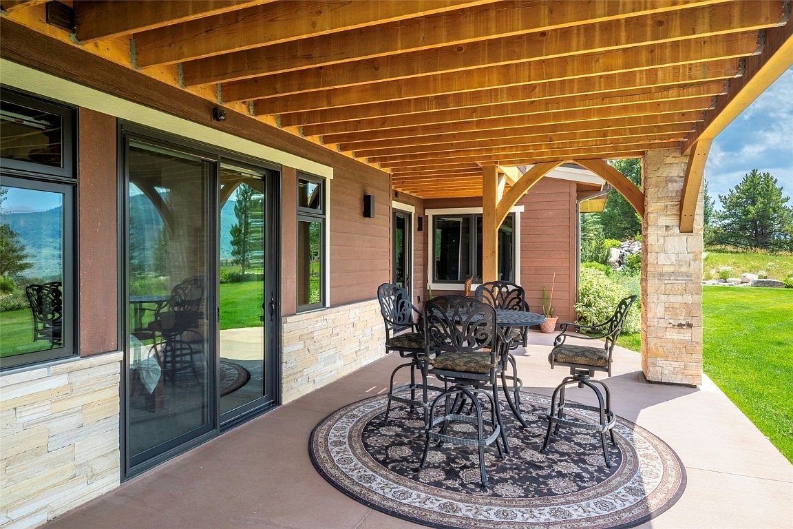 This image showcases a covered patio area, perfect for outdoor entertaining. The patio features a round outdoor rug beneath a wrought iron table and chairs set. The space is sheltered by a wooden pergola supported by stone pillars, and the patio opens onto a well-maintained lawn, creating an inviting and relaxing atmosphere.