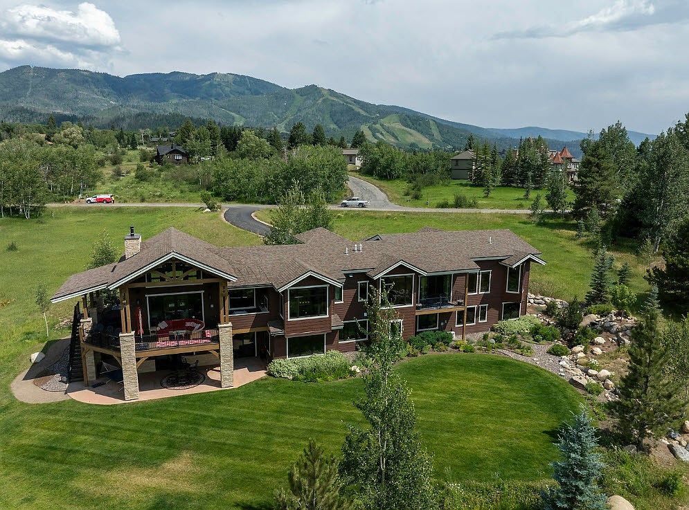 This aerial view showcases a large, luxurious home with a brown exterior and a well-manicured lawn. The house features a spacious deck, multiple windows, and is nestled in a scenic location with mountains in the background. A winding driveway leads to the property, enhancing its curb appeal and sense of privacy.