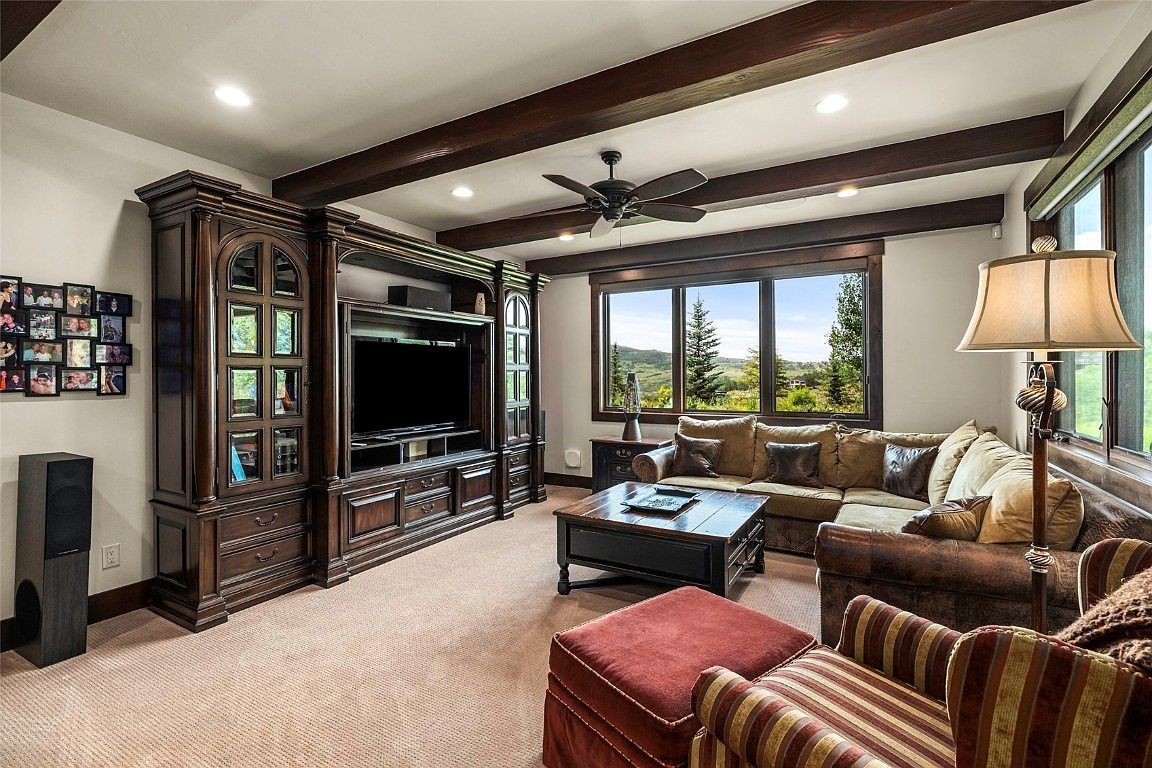 This is a cozy living room featuring a large entertainment center with a television, a comfortable sectional sofa, and an armchair with an ottoman. The room has dark wood beams on the ceiling and a large window with a view of the outdoors. The overall style is traditional and inviting, perfect for relaxing and entertaining.