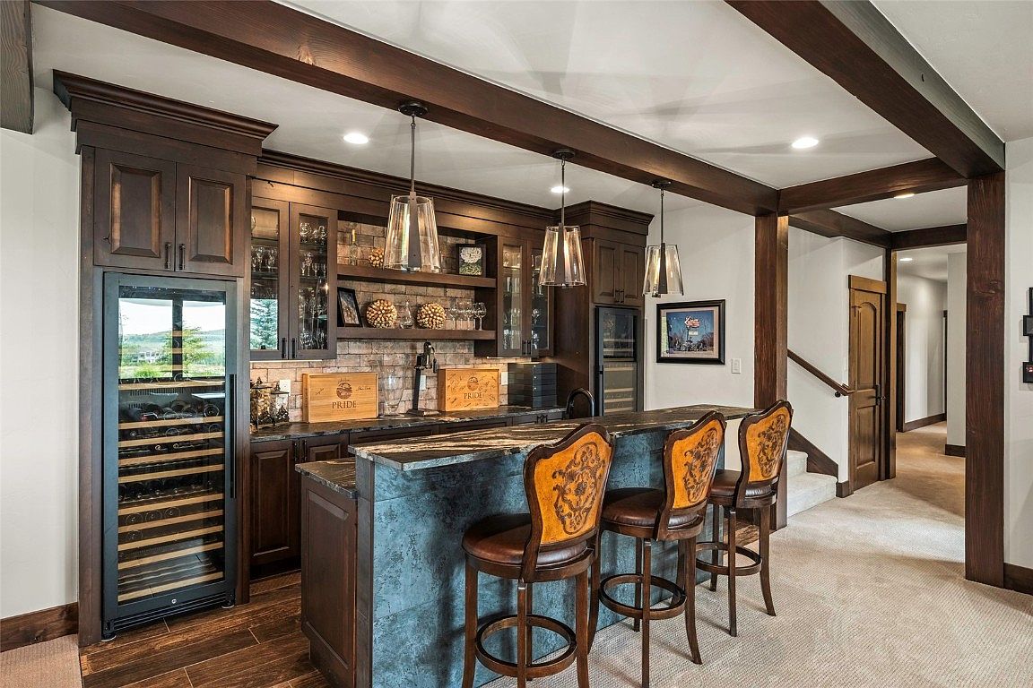 This interior shot showcases a stylish basement bar area, complete with dark wood cabinetry, a stone-faced bar, and three bar stools. Pendant lights hang above the bar, illuminating the countertop, while a wine refrigerator is built into the cabinetry. The space exudes a sophisticated and inviting atmosphere, perfect for entertaining.