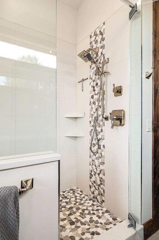 This is a well-lit bathroom featuring a modern shower with white tile walls accented by a vertical strip of pebble mosaic tiles. The shower floor is also covered in pebble mosaic tiles, complementing the shower fixtures and hardware. A glass shower door and built-in shelves add to the functionality and aesthetic appeal of the space.