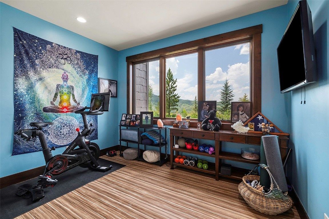 This is an interior shot of a home gym featuring a stationary bike positioned in front of a vibrant tapestry. The room is well-lit with natural light streaming through a large window, showcasing a scenic outdoor view. A console table displays decorative items and workout equipment, contributing to a functional and aesthetically pleasing space.
