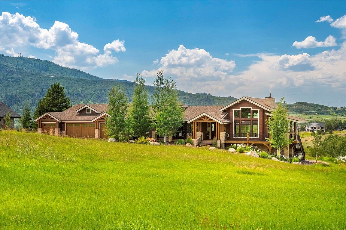 This image showcases the front exterior of a beautiful home with a well-manicured lawn. The house features a combination of wood and stone elements, a covered entryway, and large windows that provide ample natural light. The surrounding landscape includes mature trees and a mountain backdrop, enhancing the property's appeal and creating a serene atmosphere.