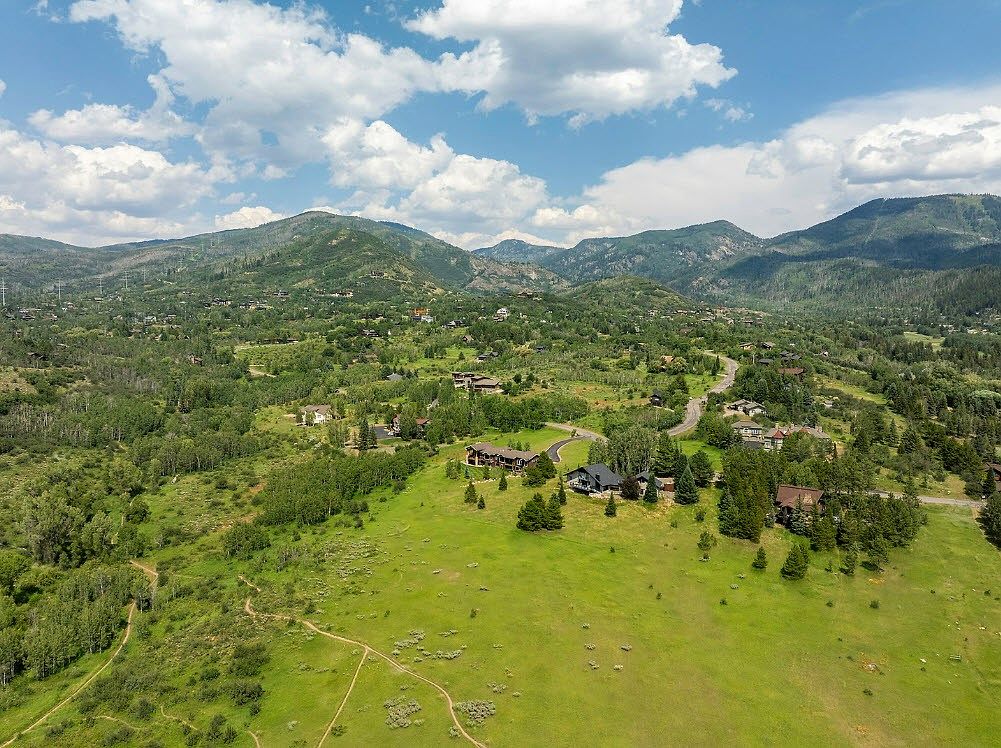 This aerial view showcases a sprawling property nestled in a mountainous landscape. The foreground features a large, well-maintained lawn with walking paths, transitioning into a wooded area with several houses scattered throughout. The background reveals rolling hills and mountains under a partly cloudy sky, creating a sense of privacy and natural beauty.