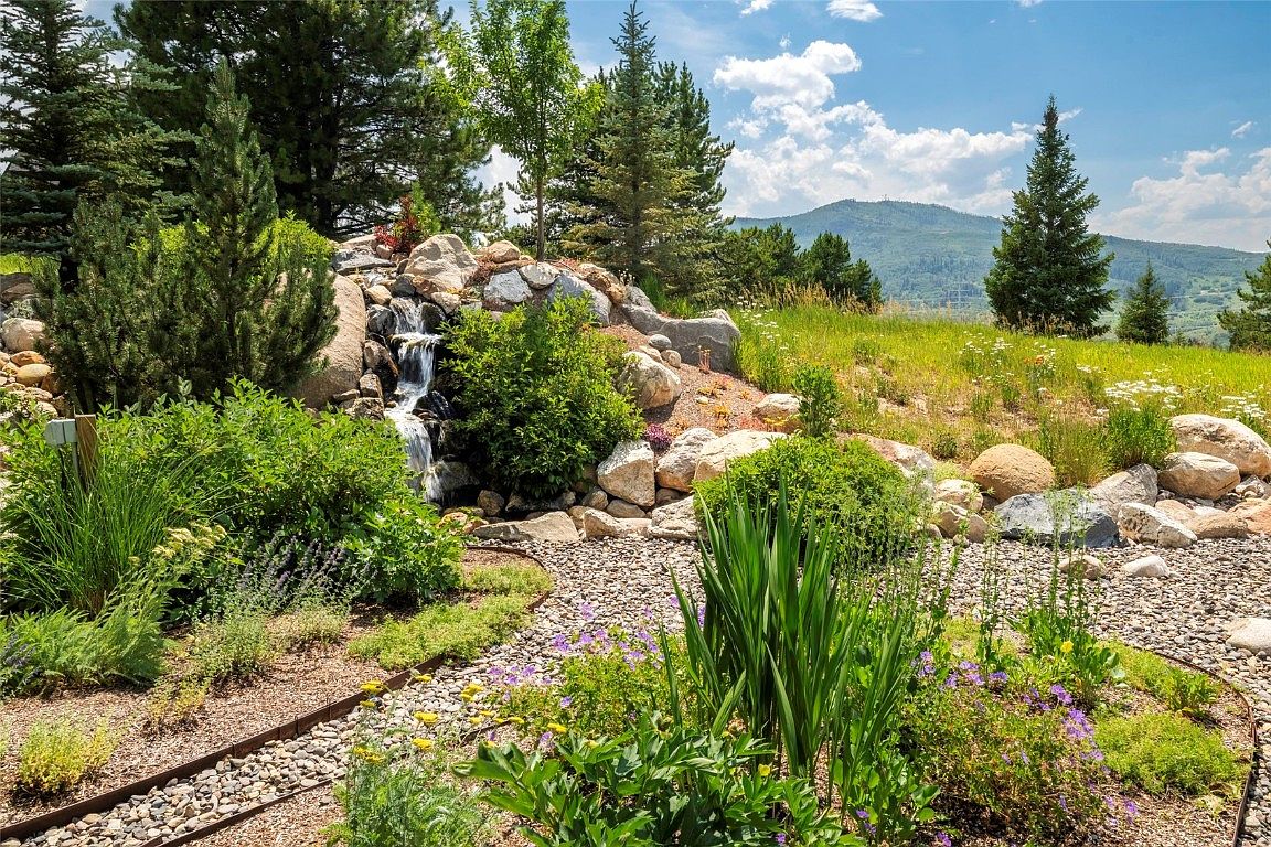 This image showcases a beautifully landscaped yard featuring a rock garden with a small waterfall. Lush greenery, including various plants and trees, surrounds the water feature, creating a serene and natural environment. The garden is complemented by a gravel pathway, and a mountain is visible in the background, adding to the scenic appeal.
