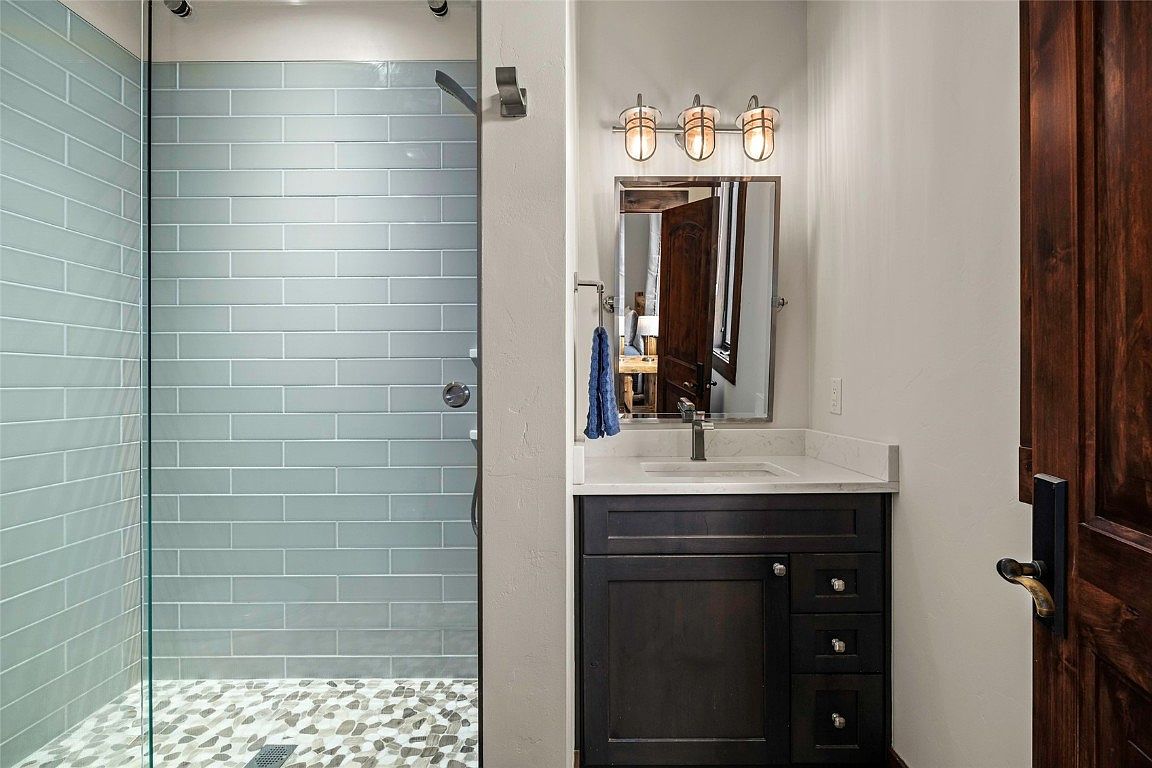 This is a well-appointed guest bathroom featuring a glass-enclosed shower with light blue subway tiles and a pebble floor. The vanity has a dark wood cabinet, a white countertop, and a modern faucet, complemented by a three-light fixture above the mirror. A dark wood door adds a touch of warmth to the space.