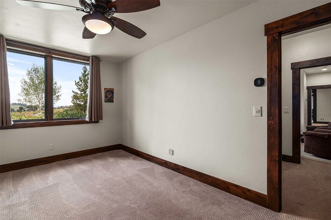 This is a well-lit bedroom featuring neutral-toned carpet and walls, complemented by dark wood trim around the baseboards, window, and doorway. A ceiling fan with a light fixture is present, and a window offers a view of the outdoors. The room appears clean and spacious, suitable for a guest bedroom.