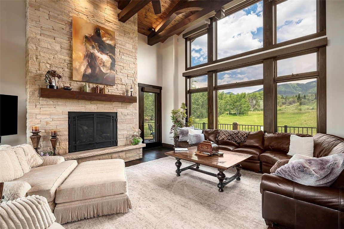 This is an interior shot of a luxurious living room featuring a stone fireplace with a wooden mantel and artwork above. Large windows offer a view of a green landscape, and a comfortable leather sectional sofa and a beige armchair with an ottoman provide ample seating. The room has a high ceiling with wooden beams, creating a spacious and inviting atmosphere.