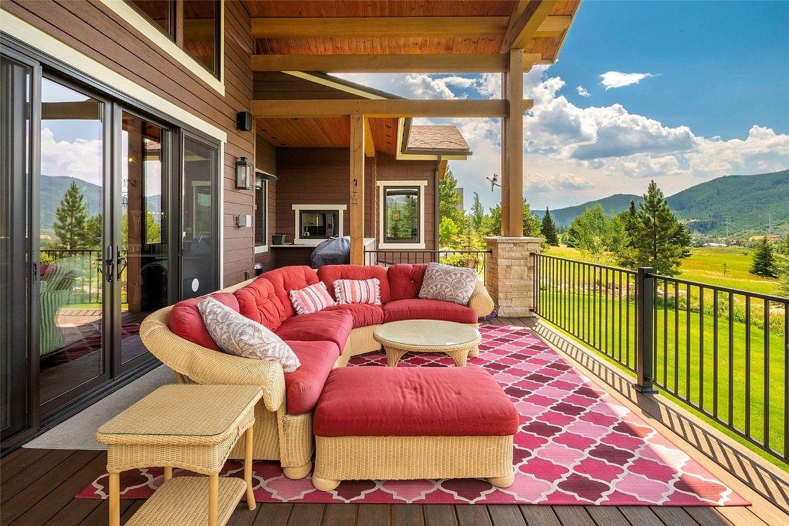 This image showcases a beautifully furnished outdoor patio or deck area. A comfortable, red cushioned sectional sofa and ottoman are arranged on a patterned rug, creating an inviting seating area. The deck features a railing with views of a lush green landscape and distant mountains, suggesting a serene and private outdoor living space.