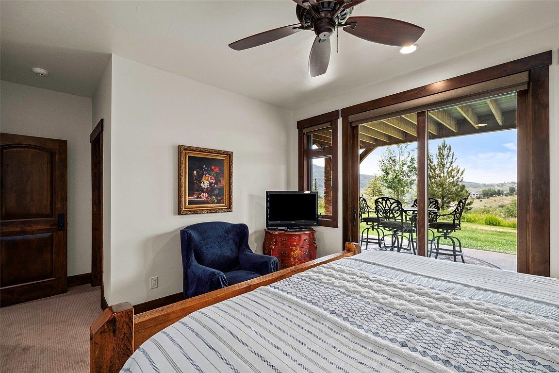 This is a primary bedroom featuring a wooden bed frame with a striped comforter, a blue armchair, and a television on a wooden chest. A painting hangs on the wall above the chair. Sliding glass doors lead to an outdoor patio with seating, offering a view of the landscape. A ceiling fan is visible above the bed.