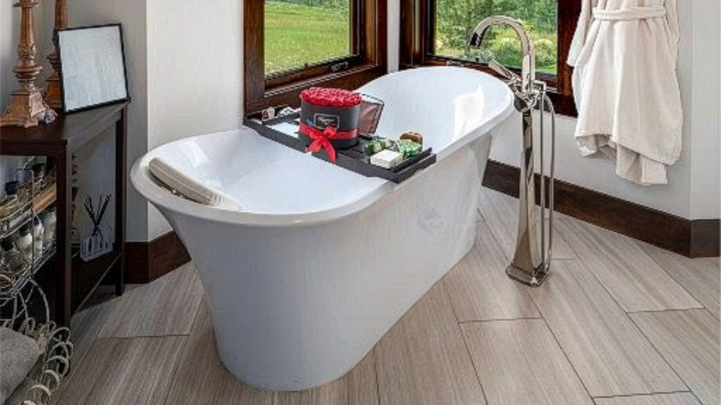 This is a well-appointed primary bathroom featuring a freestanding bathtub with a modern chrome faucet. The tub is staged with a bath tray holding flowers and toiletries. Wood-look tile flooring grounds the space, creating a spa-like atmosphere.