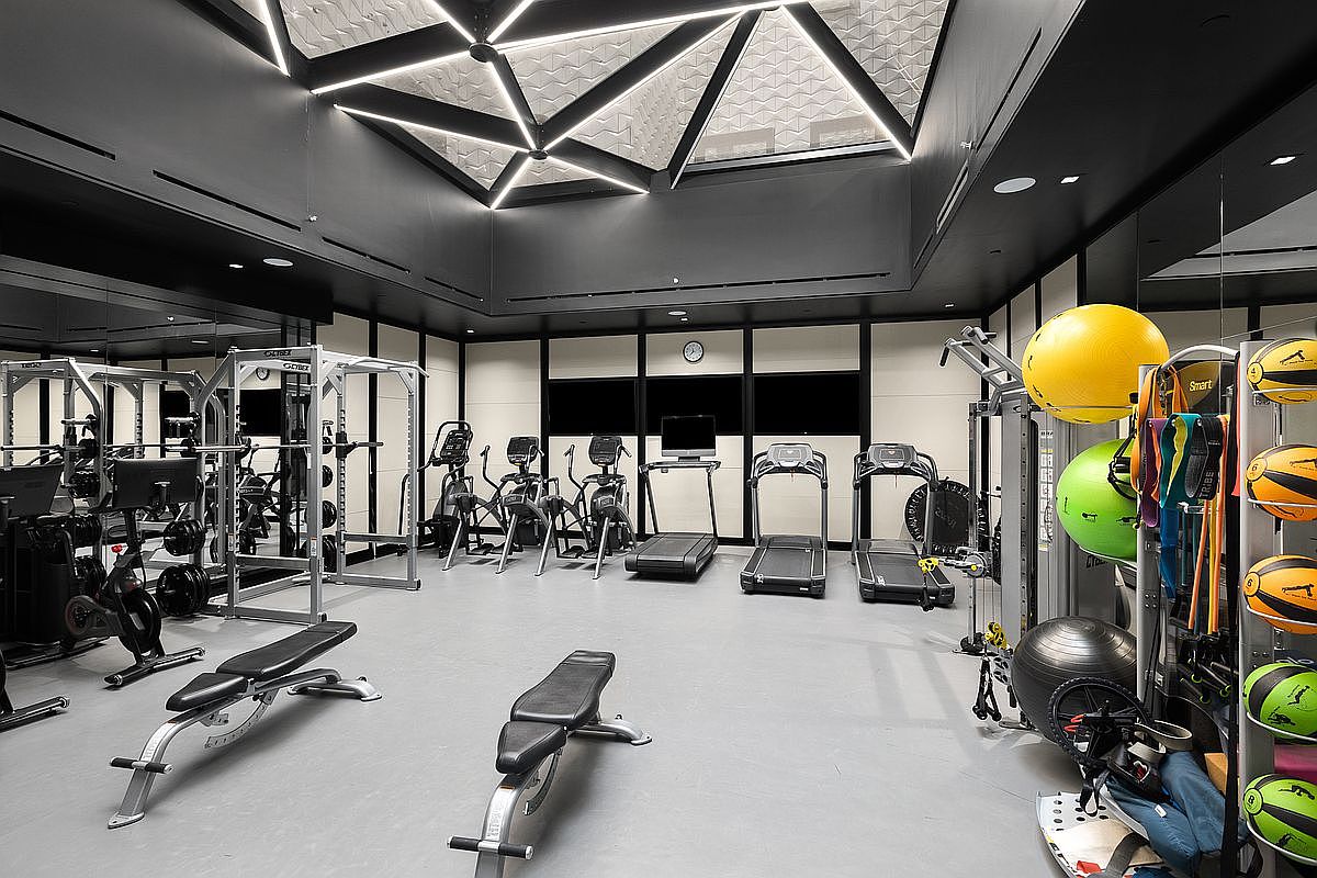 This is a well-equipped home gym featuring a variety of exercise machines, including treadmills, elliptical trainers, and weightlifting equipment. The room has a modern design with a unique geometric ceiling lighting fixture and mirrored walls, creating a spacious and motivating workout environment. The floor is a neutral gray, and the overall impression is one of a high-end, private fitness space.