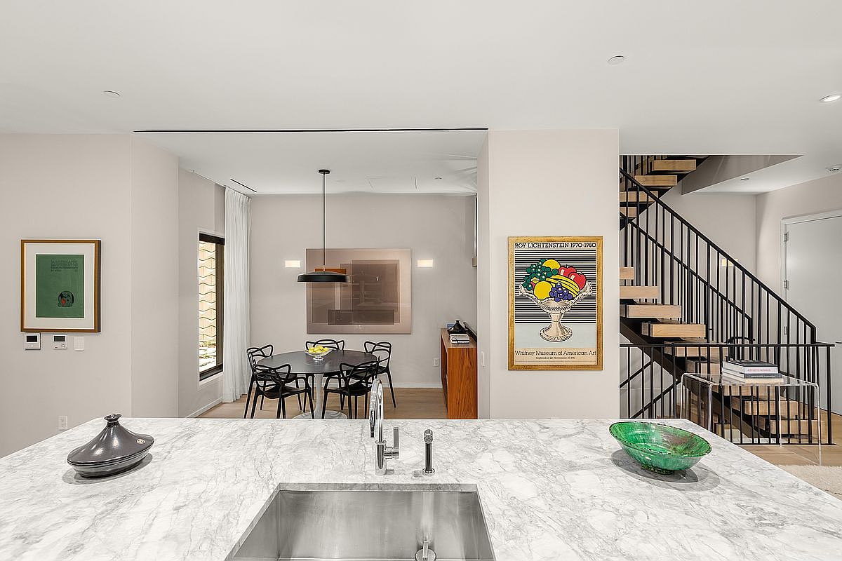 This interior shot showcases a modern kitchen with a marble countertop island and stainless steel sink. The open-concept design flows into a dining area with a round table and black chairs, while a staircase with black railings is visible on the right. Artwork adorns the walls, adding a touch of sophistication to the space.