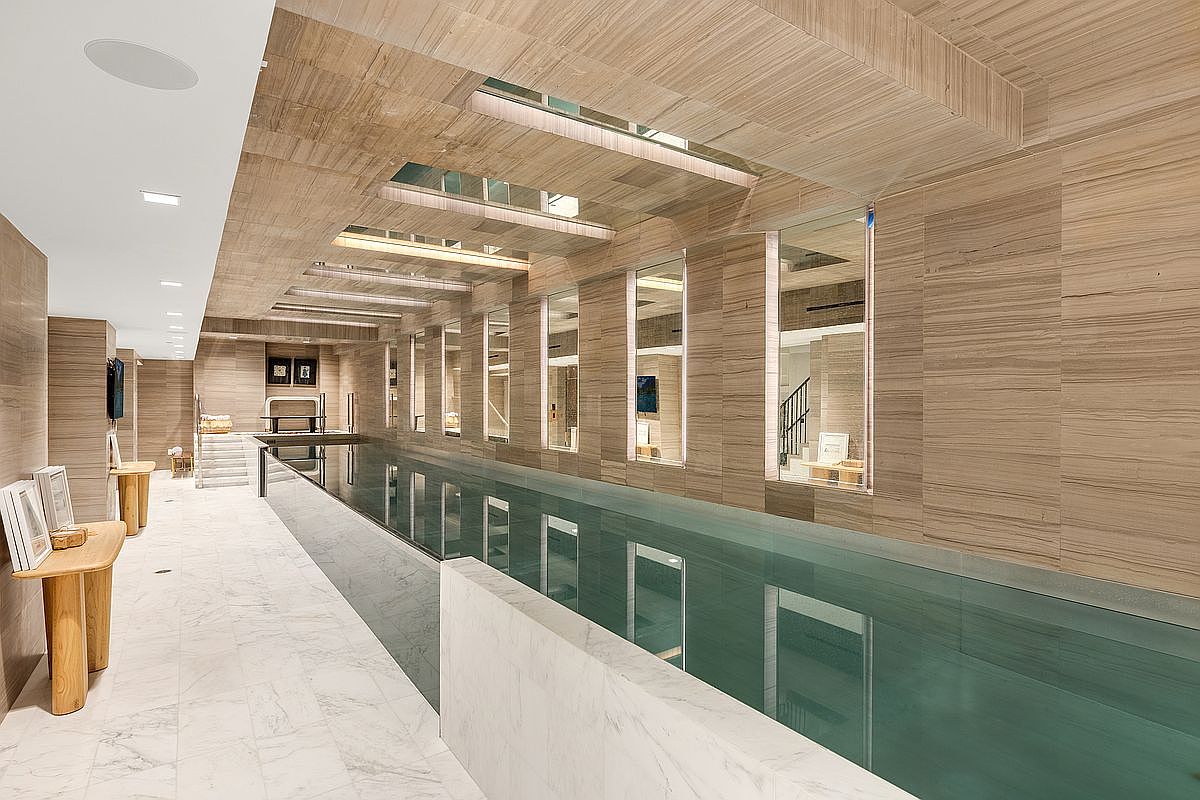 This image showcases a luxurious indoor swimming pool area, featuring sleek, light-colored wood paneling on the walls and ceiling, complemented by marble flooring. The pool itself is long and narrow, reflecting the light from the recessed windows along one side. The overall impression is one of modern elegance and tranquility, perfect for a high-end real estate listing.