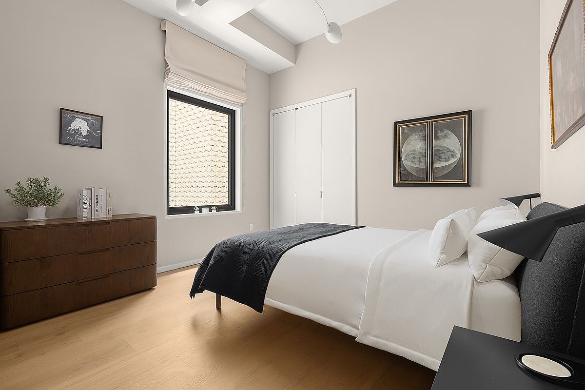 This is a well-lit primary bedroom featuring a neutral color palette, hardwood floors, and modern furnishings. A large window with a roman shade provides natural light, while artwork adorns the walls. The bed is neatly made with white linens and a dark gray throw, creating a serene and inviting atmosphere.