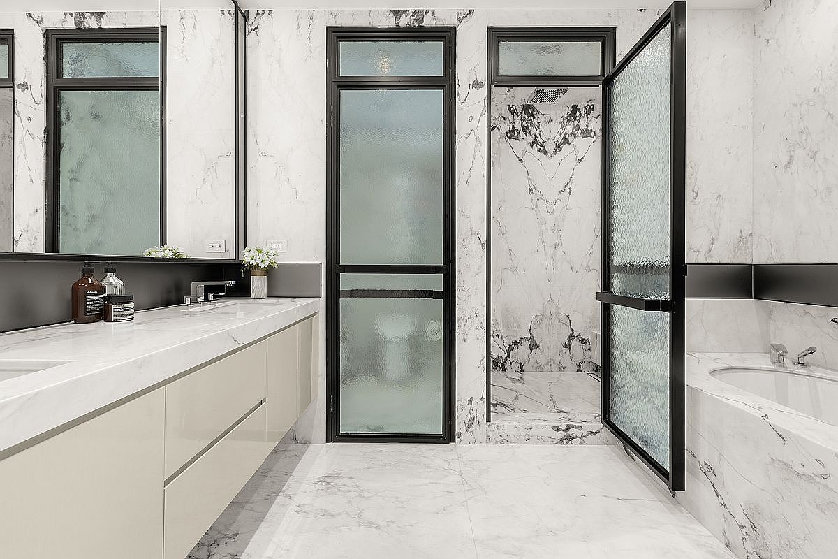 This is a luxurious primary bathroom featuring marble walls and flooring, creating a bright and elegant space. The bathroom includes a long vanity with modern fixtures, a glass-enclosed shower, and a soaking tub. Black framed windows and accents add a contemporary touch to the classic marble design.