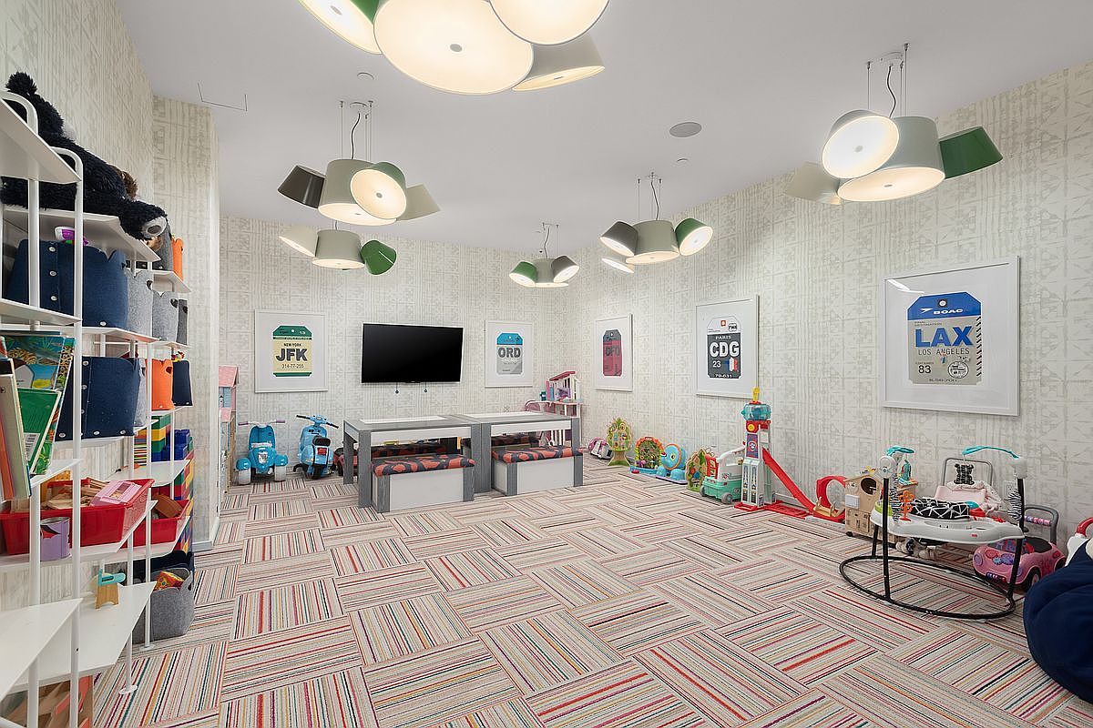 This is a well-organized and playful children's playroom featuring a striped carpet, whimsical lighting fixtures, and framed artwork of airport codes. The room includes a shelving unit filled with toys and books, a table with seating, and various play items scattered throughout, creating a fun and inviting space.