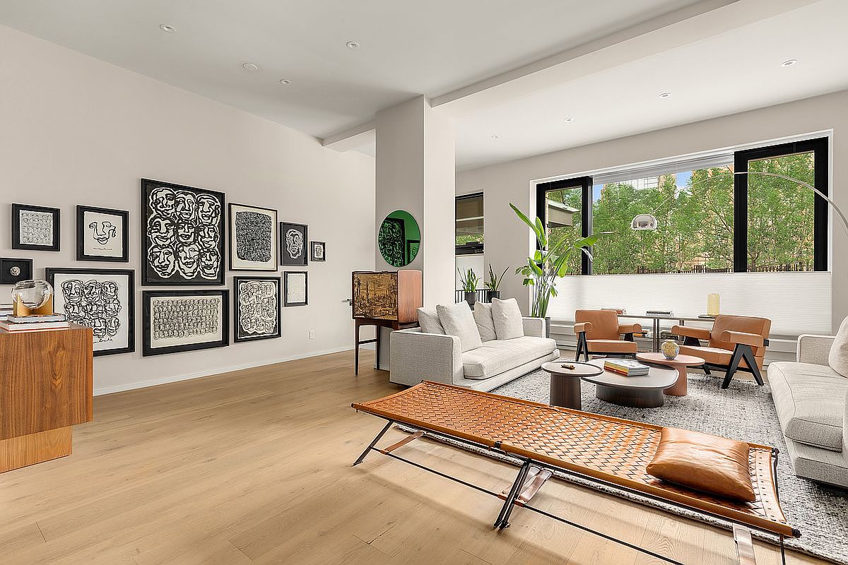 This is an interior shot of a modern living room. The room features light wood flooring, a gallery wall with framed artwork, and contemporary furniture including sofas, armchairs, and a unique woven daybed. Large windows provide natural light and views of the outdoors, creating a bright and inviting space.