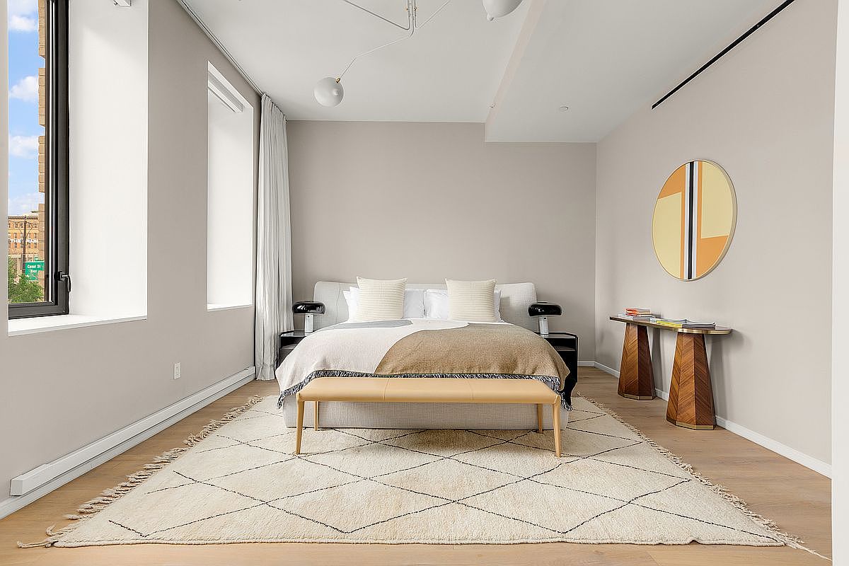 This is a well-lit primary bedroom featuring a neutral color palette. The room includes a bed with a light-colored headboard, two nightstands with lamps, and a bench at the foot of the bed. A geometric rug adds texture to the wooden floor, and a round piece of art hangs on the wall above a console table, creating a serene and stylish atmosphere.