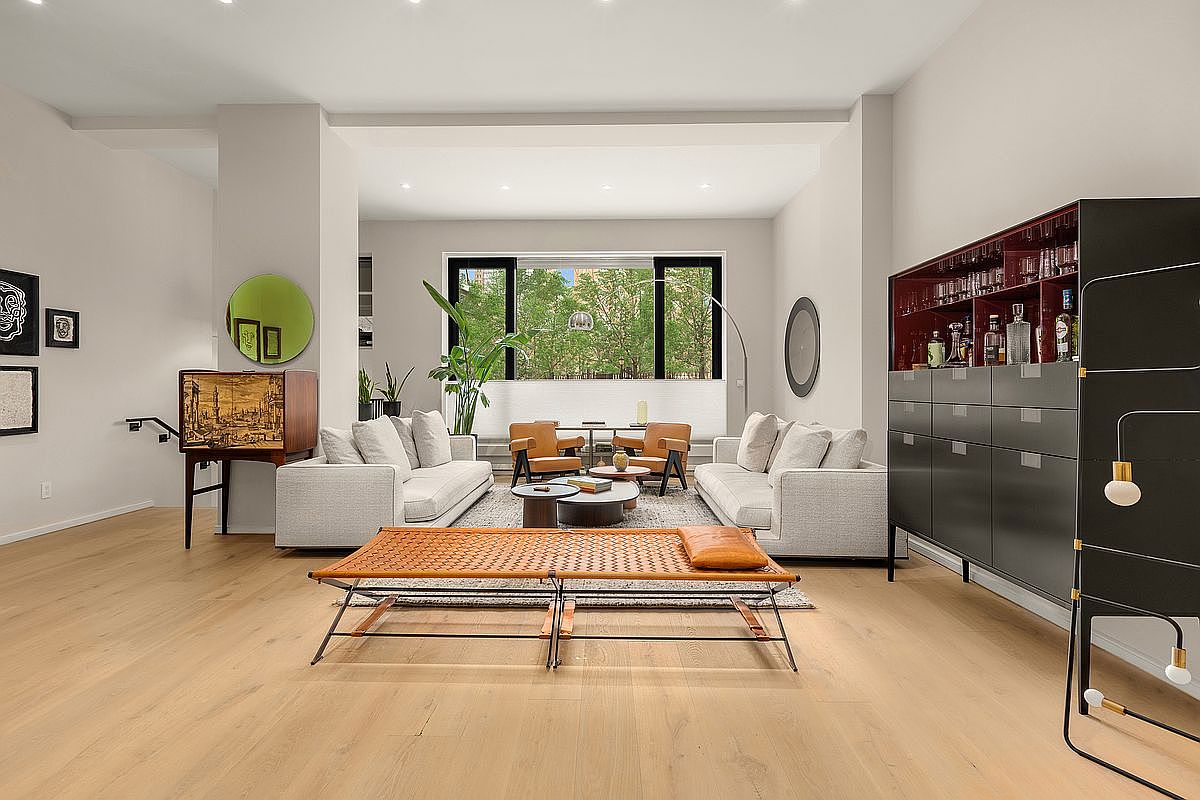 This is a spacious and modern living room featuring light hardwood floors, neutral-toned sofas, and a large window providing ample natural light. The room is stylishly furnished with a unique woven daybed, a dark bar cabinet, and various decorative elements, creating a sophisticated and inviting atmosphere. The perspective is a wide shot, showcasing the entire living space and its layout.