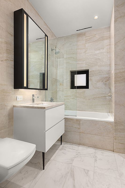 This is a well-lit primary bathroom featuring a modern vanity with a white cabinet and a light countertop, complemented by a black-framed mirror with integrated lighting. The bathroom includes a bathtub with a glass partition and a tiled wall niche, all finished with neutral-toned tiles. The flooring is a light marble, contributing to a clean and sophisticated aesthetic.