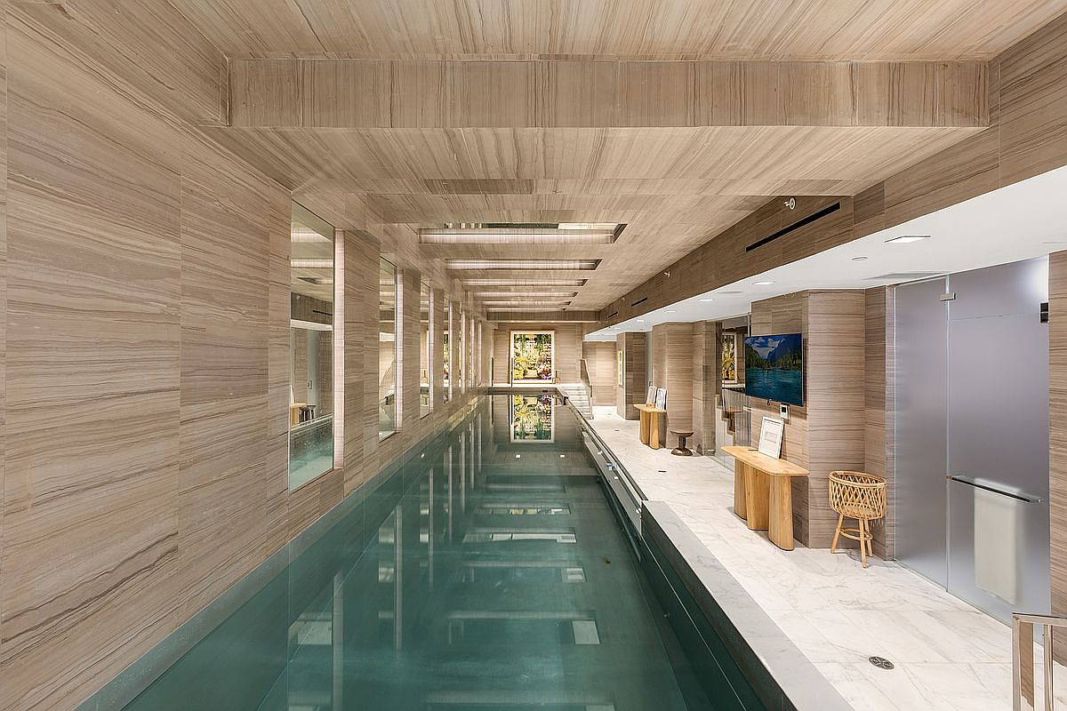 This image showcases a luxurious indoor swimming pool area. The pool is long and rectangular, with clear blue water reflecting the overhead lighting and wall mirrors. The surrounding walls and ceiling are clad in light-colored wood paneling, creating a warm and sophisticated ambiance. The space also features lounge areas with wooden tables and a wicker chair, enhancing the spa-like atmosphere.