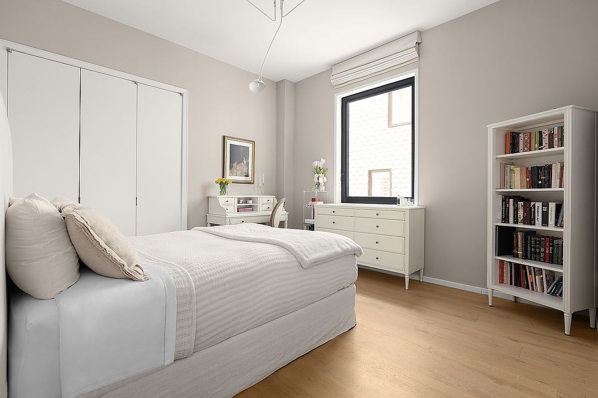 This is a bright and airy bedroom featuring a neutral color palette. The room includes a bed with white linens, a white dresser, a desk, and a bookshelf filled with books. Natural light streams in through a window, creating a serene and inviting atmosphere.
