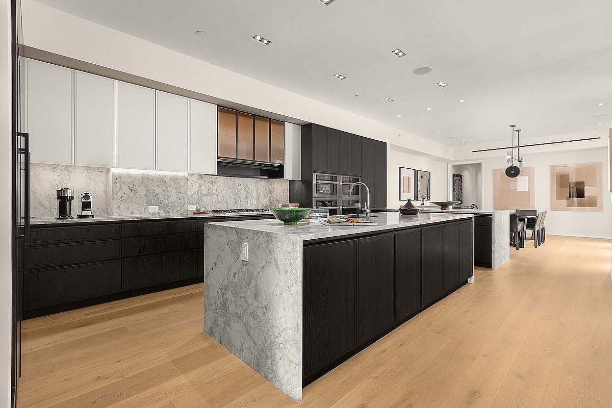 This is a modern kitchen with a large island featuring a waterfall countertop. The cabinetry is a mix of dark wood and white, with stainless steel appliances and a marble backsplash. The kitchen also features a dining area with modern lighting and artwork, creating a luxurious and functional space.