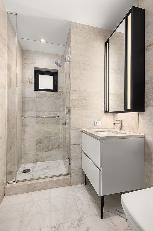 This is a well-lit primary bathroom featuring a glass-enclosed shower with a black-framed window, a modern white vanity with a light countertop, and a black-framed mirror with integrated lighting. The walls and floors are covered in light-colored marble tiles, creating a clean and luxurious aesthetic. The perspective is from the doorway, showcasing the shower and vanity area.