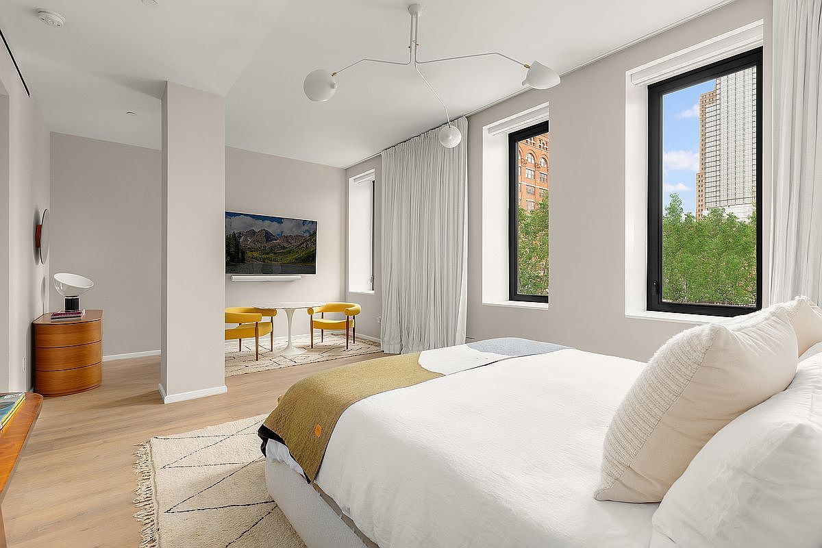 This is a bright and airy primary bedroom featuring a large bed with white linens and decorative pillows. The room has two large windows with black frames and white curtains, offering views of the city and greenery. The light hardwood floors and neutral wall colors create a serene and inviting atmosphere, complemented by modern lighting and minimalist decor.