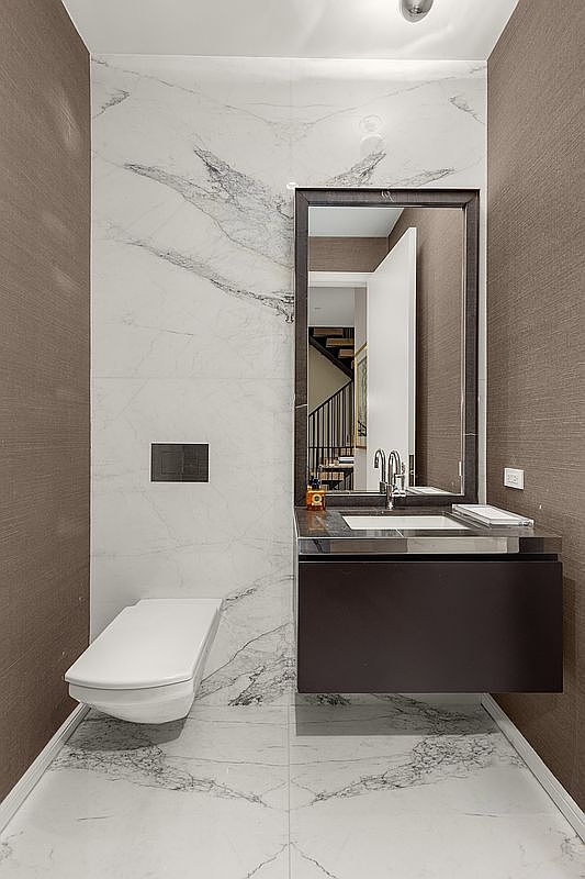 This is a modern guest bathroom featuring a wall-mounted toilet and a floating vanity with a dark finish. The walls are covered in a combination of marble-patterned tile and textured wallpaper, creating a sophisticated look. A mirror reflects a glimpse of the home's interior, adding depth to the space.