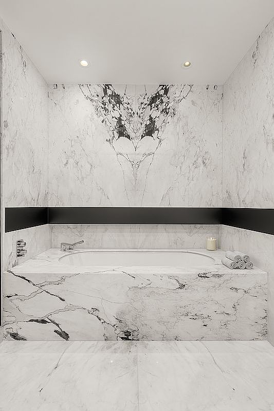 This is a luxurious primary bathroom featuring a marble-clad bathtub with a black accent stripe. The walls and floor are also covered in marble, creating a seamless and elegant look. The bathtub is surrounded by a marble deck, and there are rolled towels and a candle on the side.
